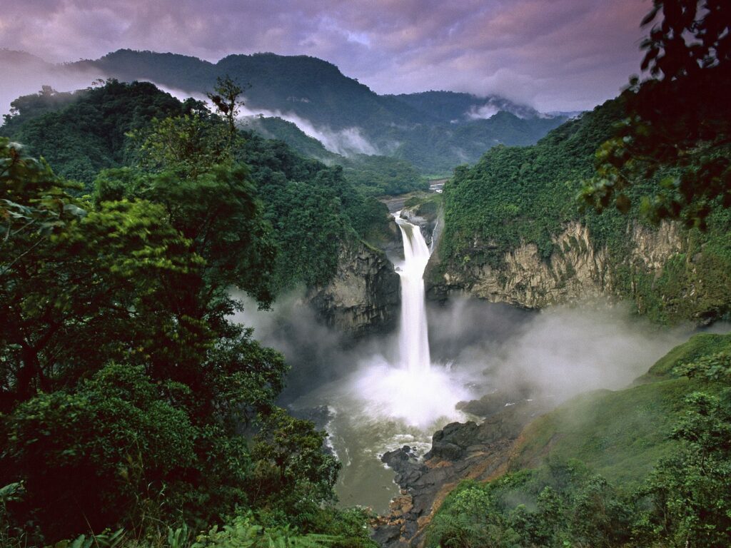 Waterfall dropping over a cliff into a pool in the middle of a hilly rainforest.