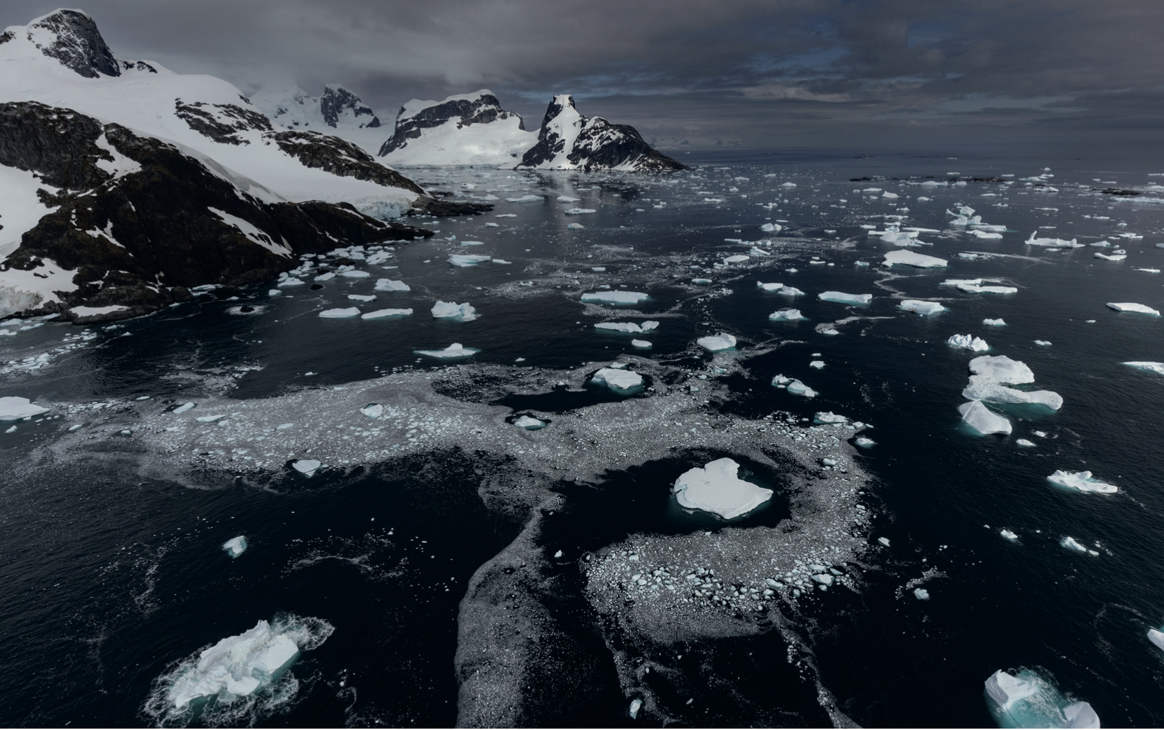 Aerial photograph of the sea withicebergs scattered over it and snowy mountains in the distance taken from a helicopter flying over Cape Tuxen, Antarctica.