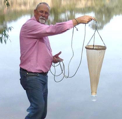 David Seamer stood on a lakes edge in a pink shirt and blue trousers collecting samples while holding a conical net over the water.