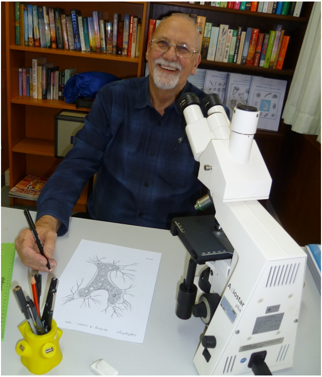 David Seamer looking at the camera smiling while sat at a table with a pencil in his hand and a large white microscope on his left and a partially complete drawing of a microorganism on his right.