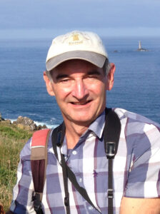 Photograph of Mike Langman, nature illustrator, on a coastal path wearing a checked shirt, cap and with a camera around his neck.