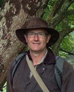 Robert Wolton portrait, showing him from the chest upwards, stood wearing a brown hat, coat and bag with an old tree in the background.