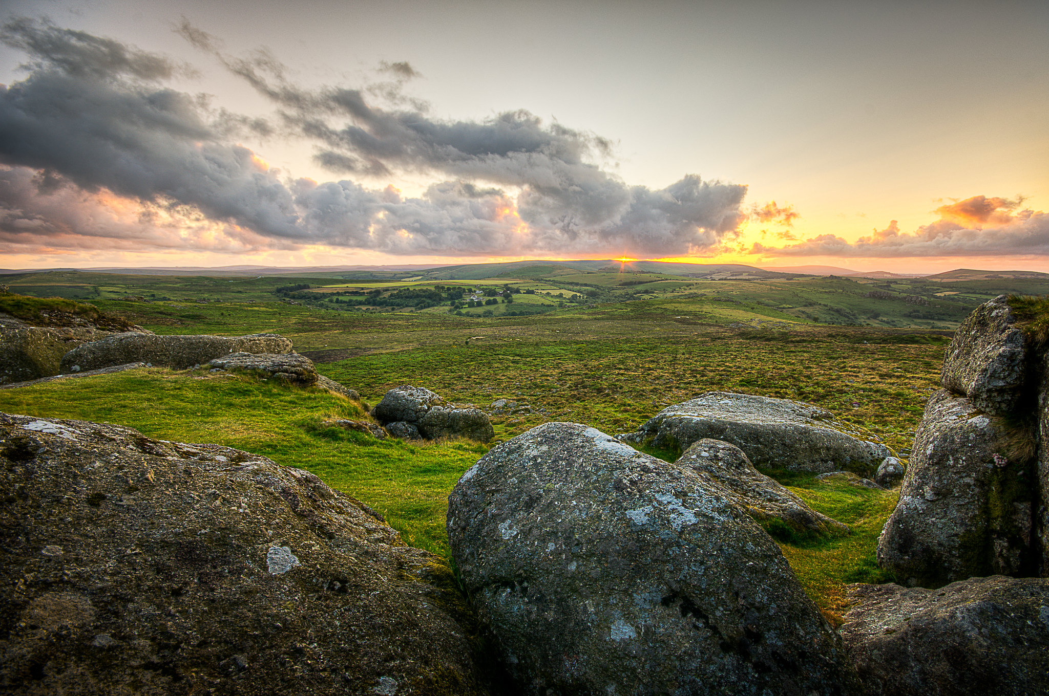 Dartmoor sunset taken from behind some rocks looking over Dartmoor's heathland with clouds in the sky and the sun setting behind them.