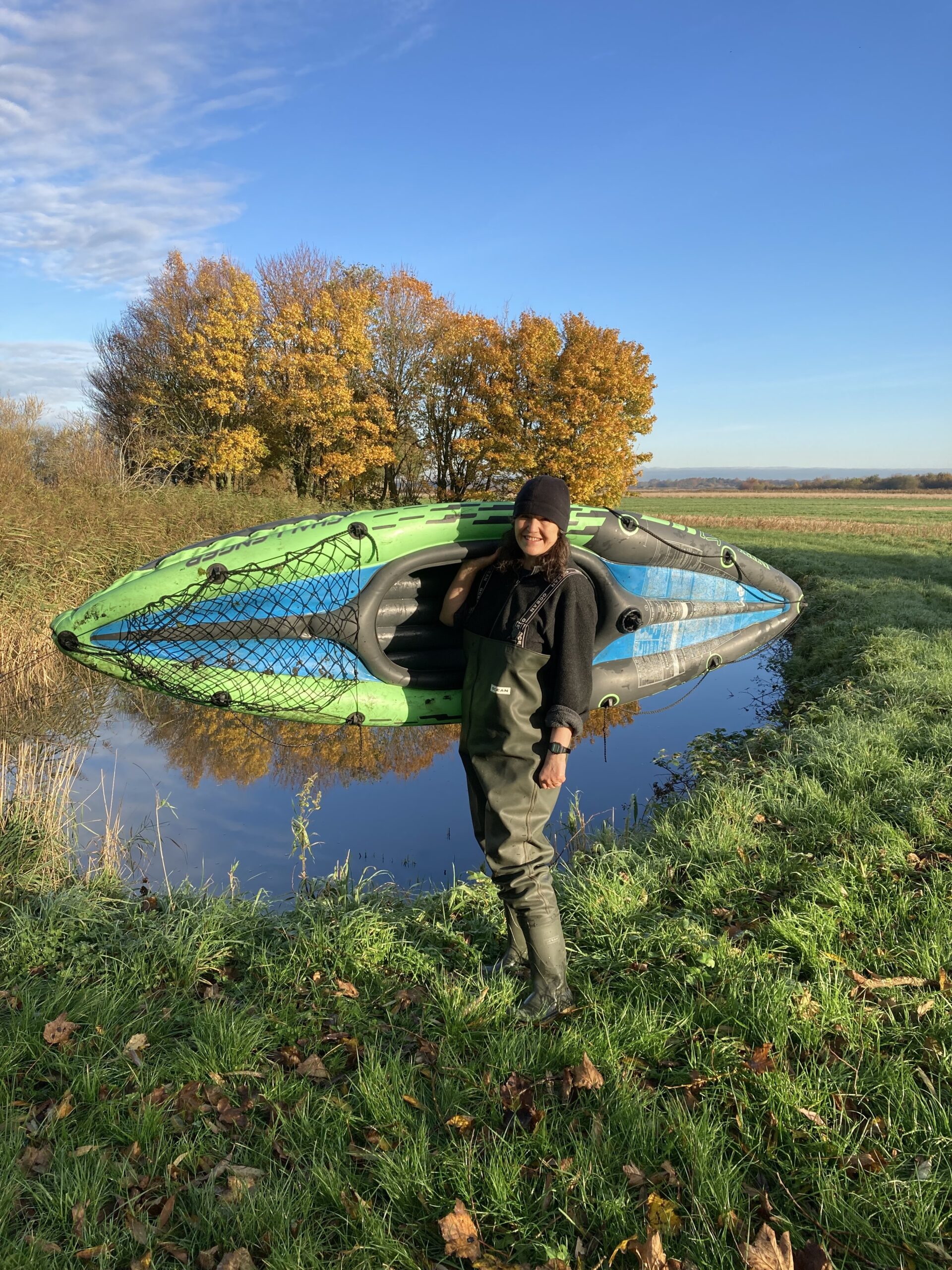 Penny Williams carrying an inflatable kayak by a pond on a sunny day with blue skies.