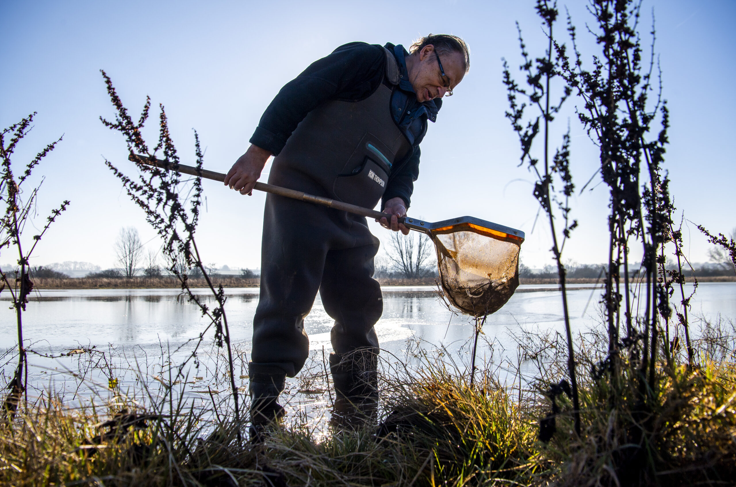 Jeremy Biggs wearing wellies on the side of a large pond looking at something he has caught in a net on a sunny day with clear, blue skies.