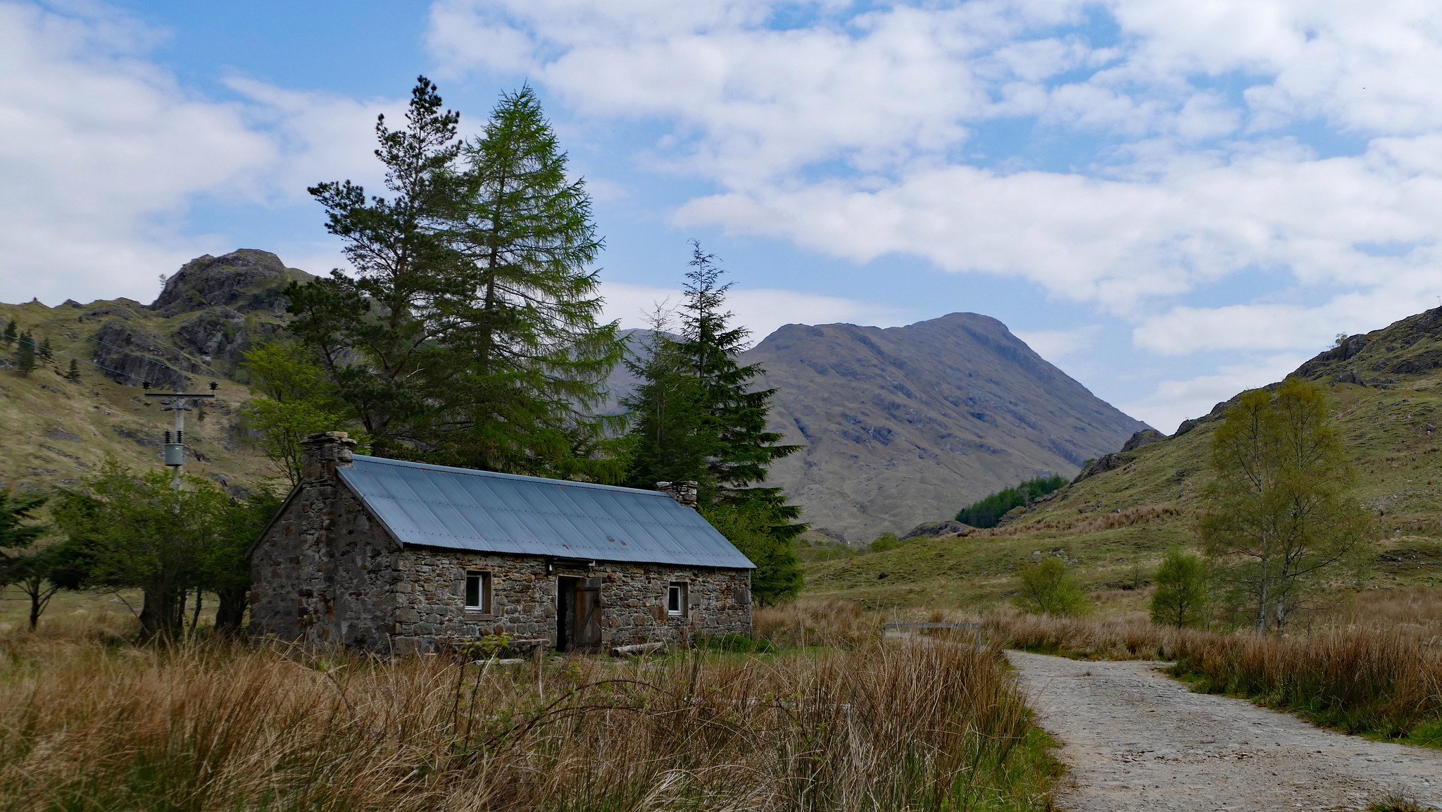 A bothy nestled under some large evergreen trees on the side of a stony track going towards the Scottish mountains in the background.