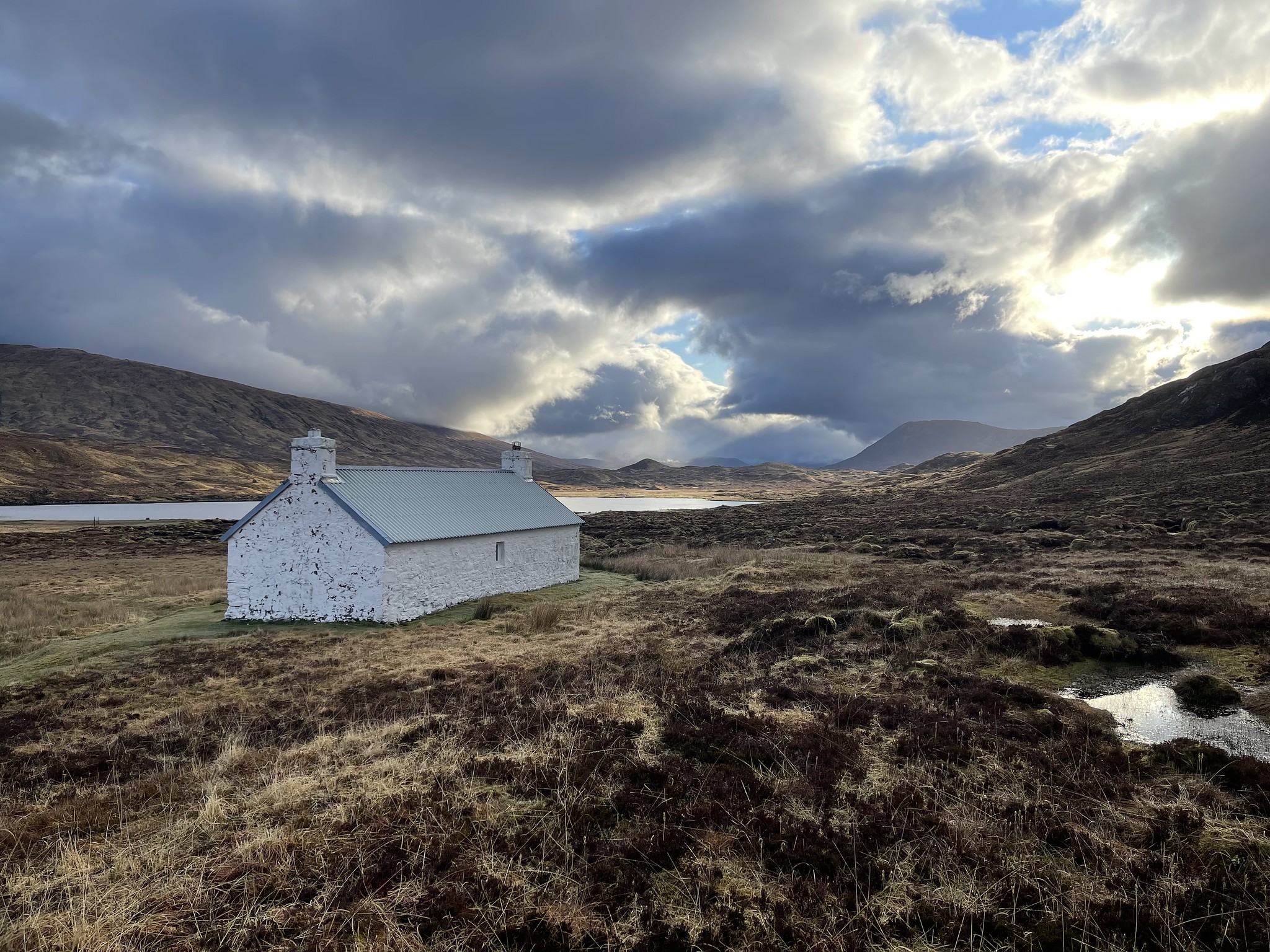 White washed, stone bothy near a river or lake one some land covered in brown grass and gorse, with Scottish mountains in the far distance.