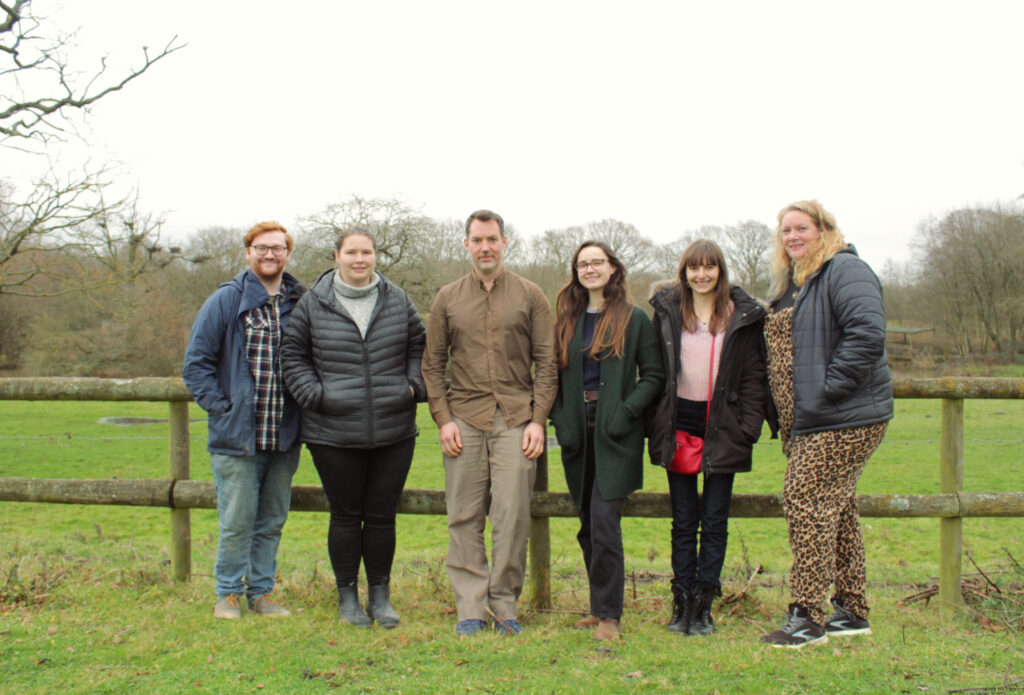 Six people smiling in front of a wooden fence in a field.