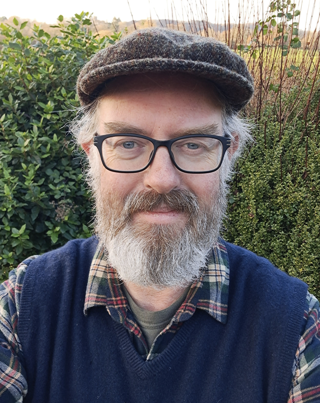 Portrait of author Christopher Hart wearing a checked shirt, gillet and flat cap with a large hedge behind him.