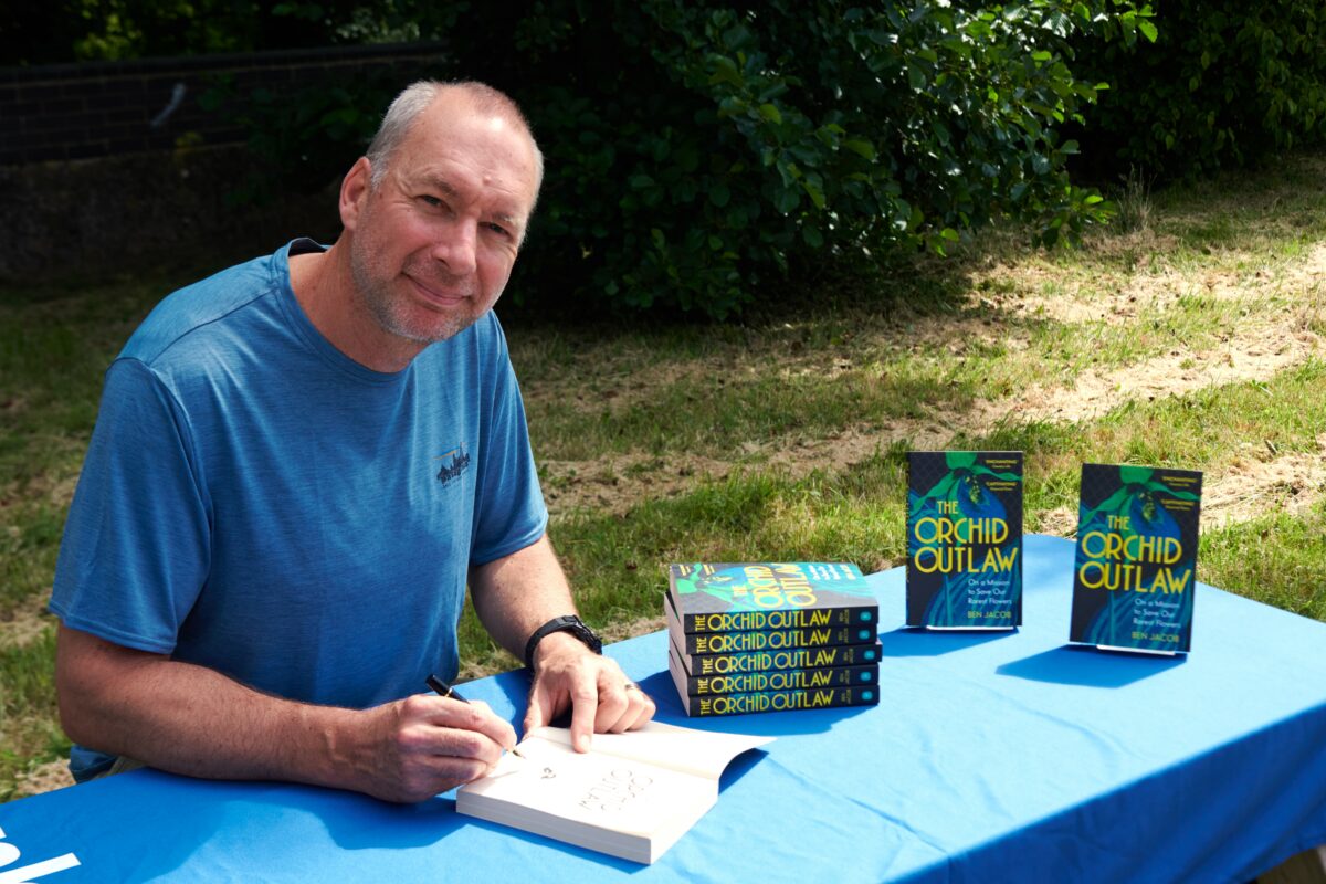 Ben Jacob, in a blue t-shirt, signing his book The Orchid Outlaw sat on a bench with grass and trees behind him.