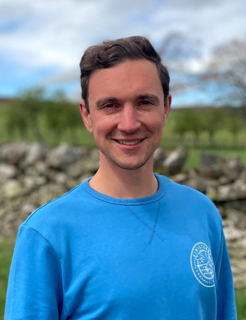 A headshot of a man in a blue tshirt smiling at the camera. 