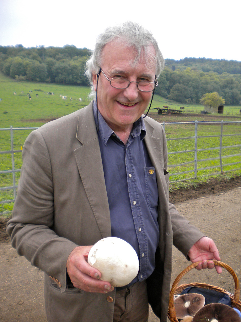 Richard Fortey in a jacket and shirt with a wicker basket full of large brown mushrooms and a white puffball mushroom in his hand.