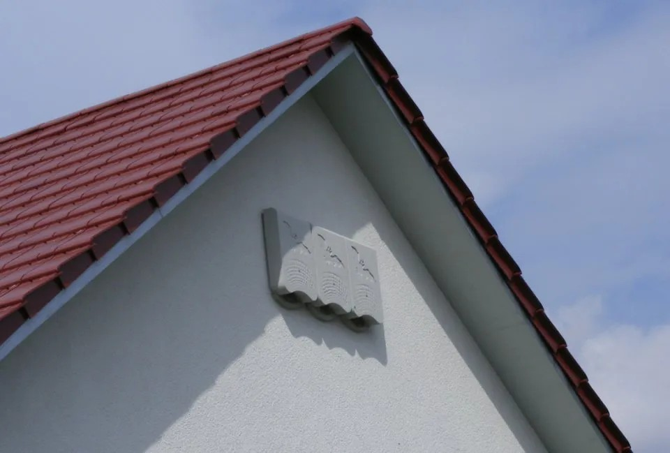 A three-part bat box located under the gables of a house, it is grey and has entrance holes at the bottom of the box