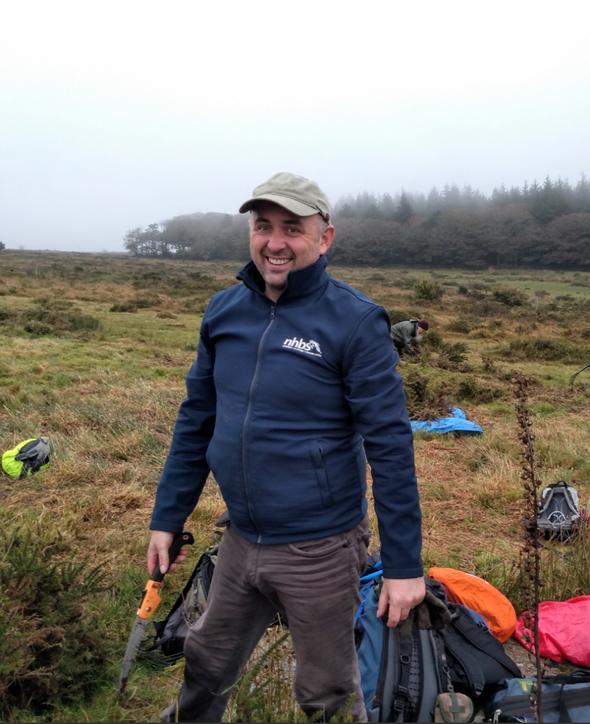 Dan is wearing a blue jacket and a cap, he is standing in the middle of grassland with forest in the background and is smiling for the camera